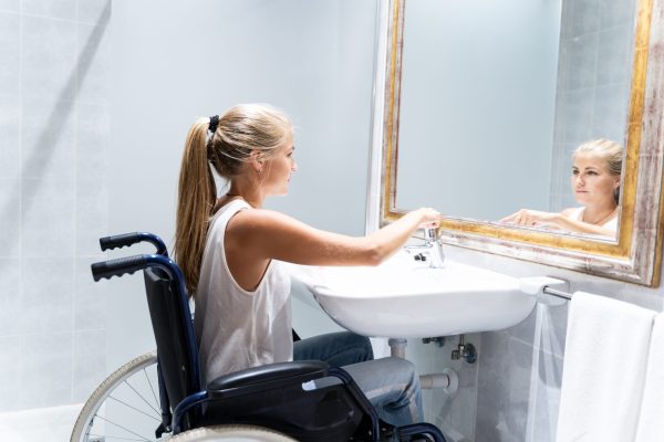 Blonde girl in wheelchair with long hair turning on the water tap in a bathroom in front of the mirror
